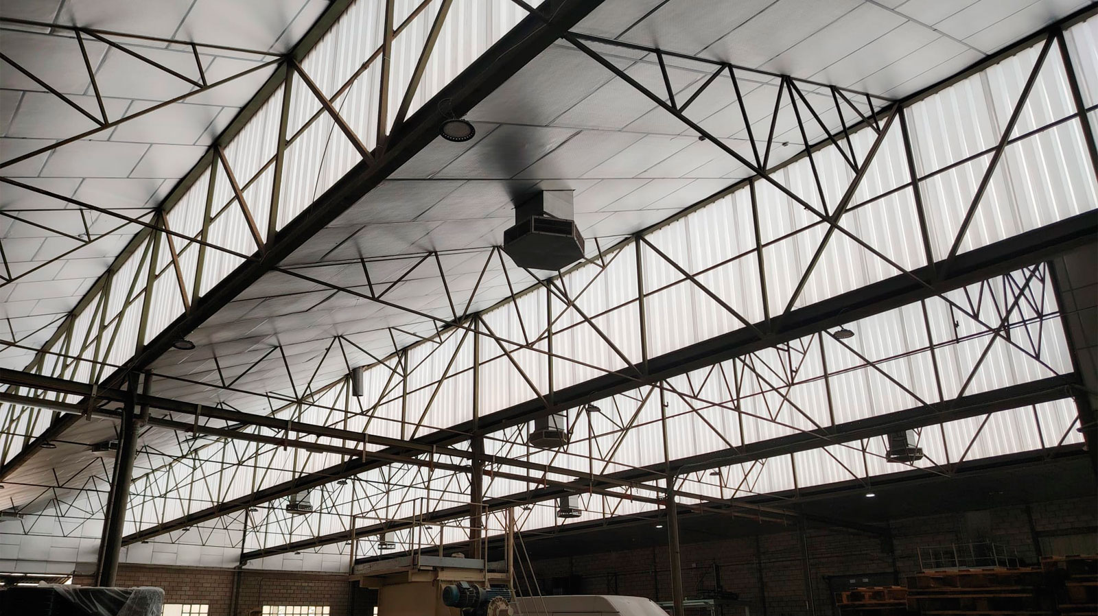 Interior de nave industrial con cubierta metálica, estructura de cerchas de acero y equipos de climatización suspendidos del techo.